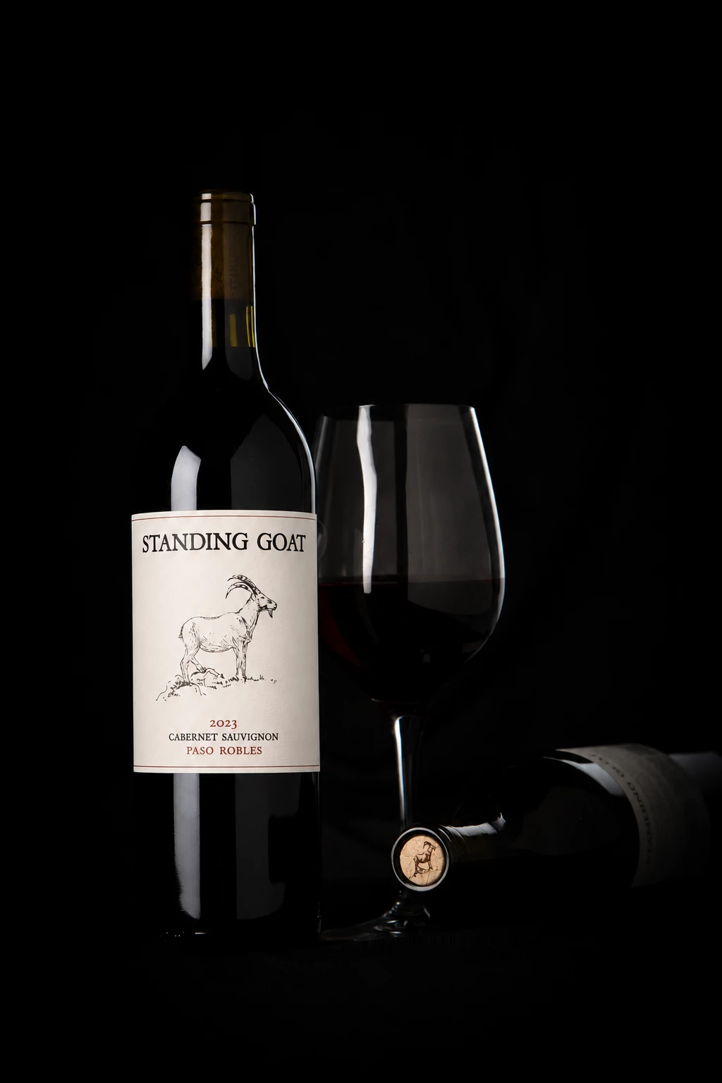 Standing Goat Cabernet Sauvignon beside a glass of red wine in a dark studio setting